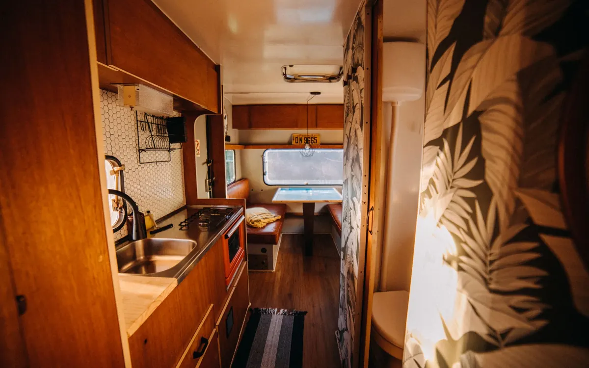 The inside of a camper with a wooden kitchenette, a small sink and cooktop, warm lighting and a dining nook by the window at the far end.