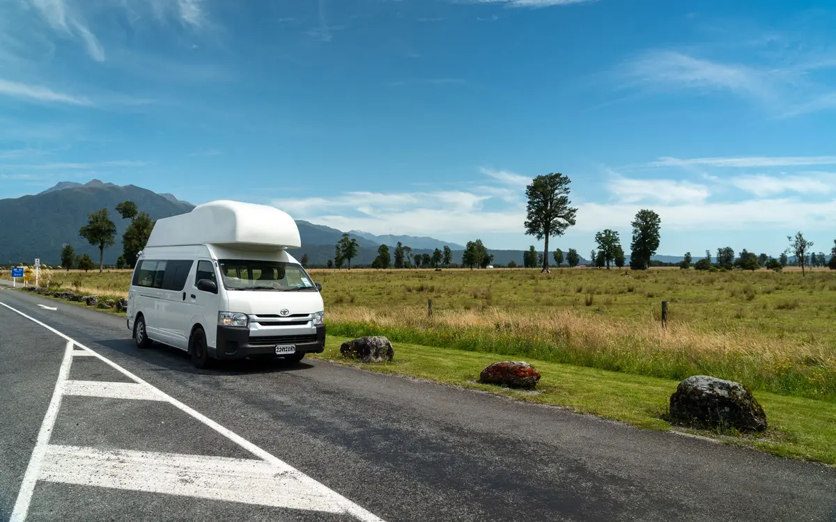 A white high roof campervan parked on the shoulder of an empty country road, with open grassland, scattered trees and blue mountains behind it.