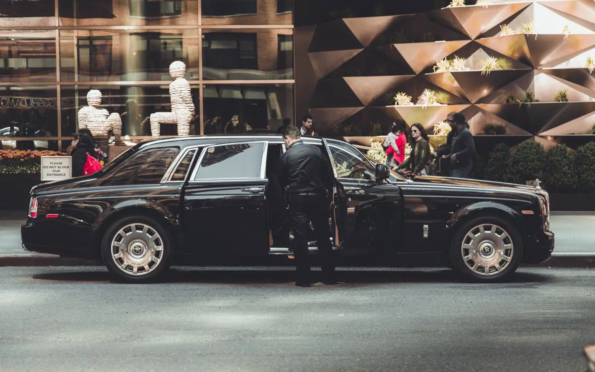 A black Rolls Royce Phantom at the kerb on a New York City street while a driver in a dark suit holds the rear door open.