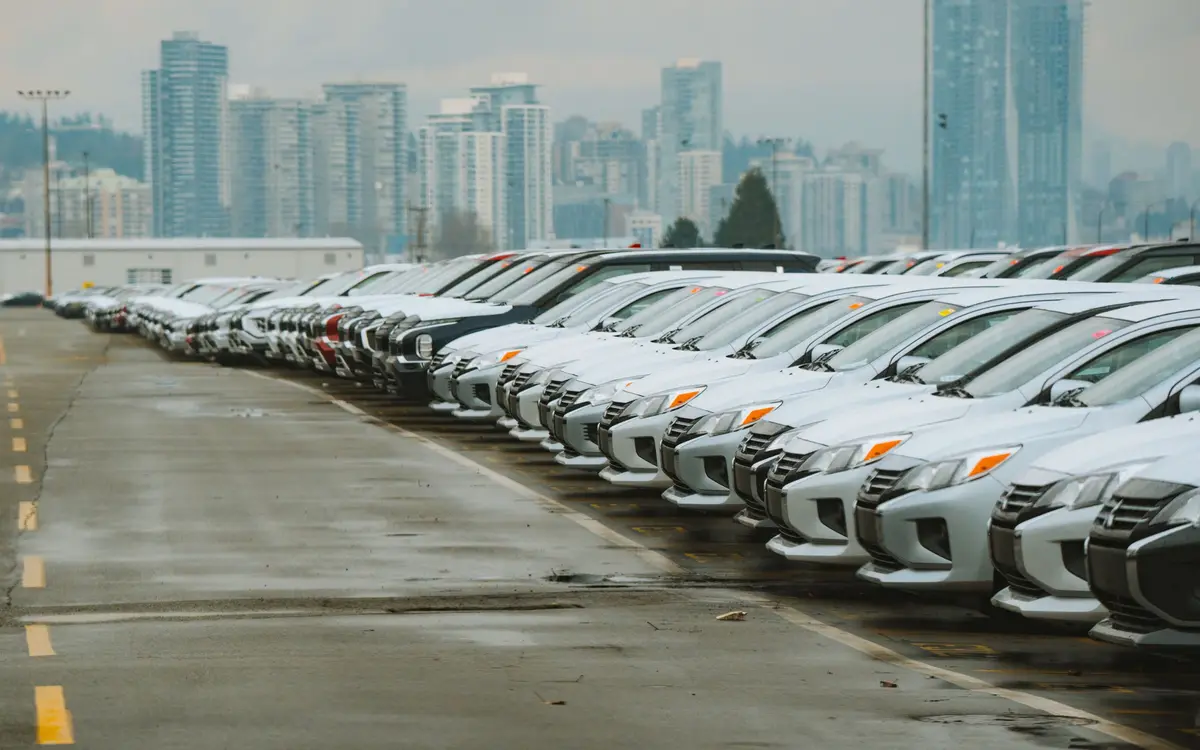 A long row of white and silver cars parked nose out on a wet asphalt lot, with city tower blocks in the haze behind them.