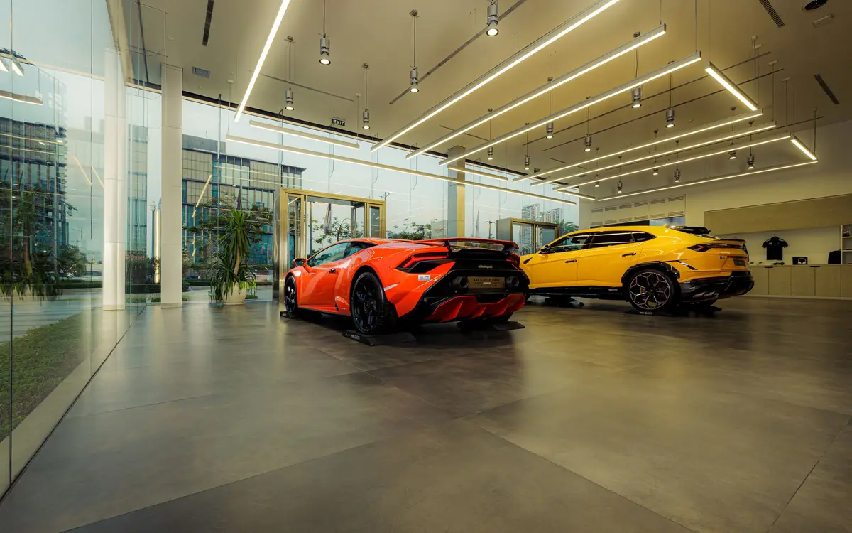 An orange Lamborghini Huracan and a yellow Lamborghini Urus parked on the polished floor of a glass fronted showroom lit by ceiling strip lights.