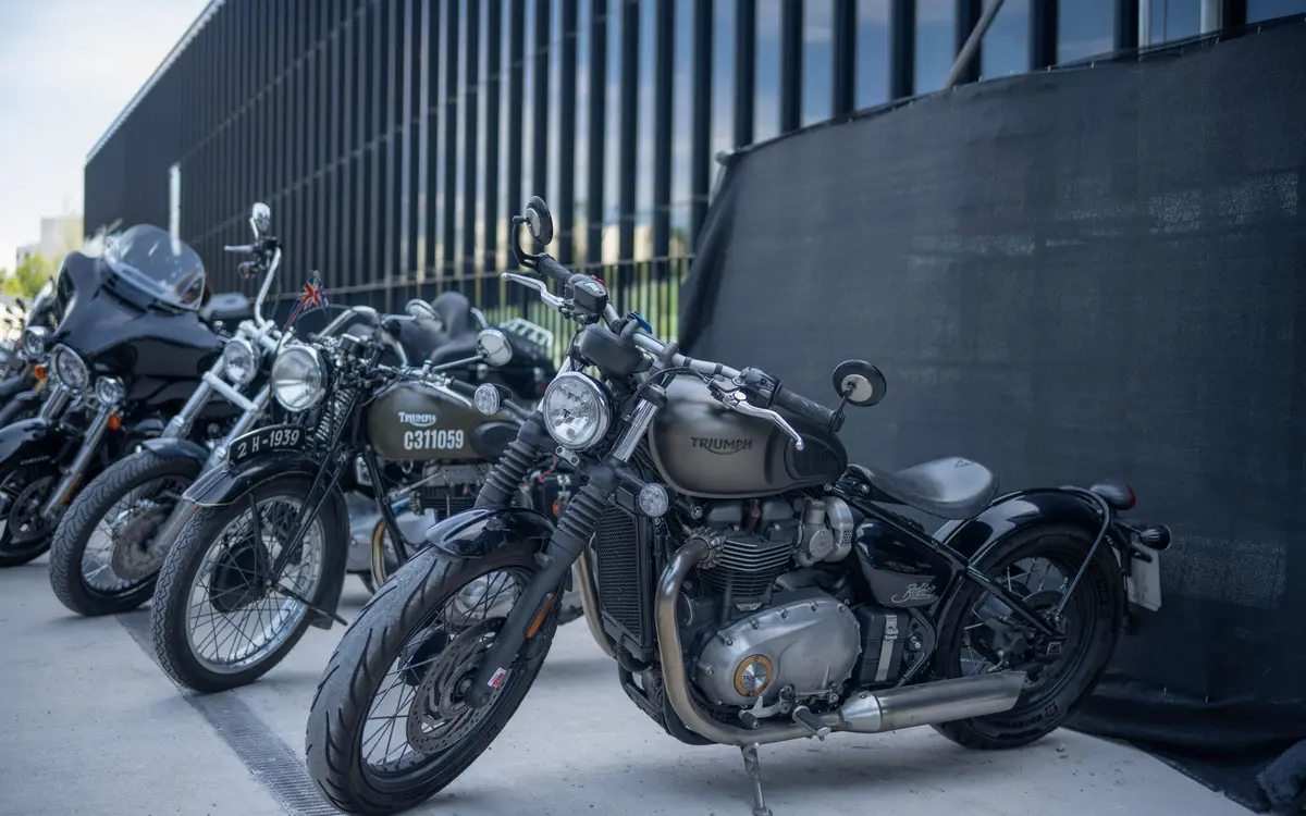 A row of Triumph motorcycles, including a bobber and a vintage machine, parked at an angle on concrete beside a dark slatted building.