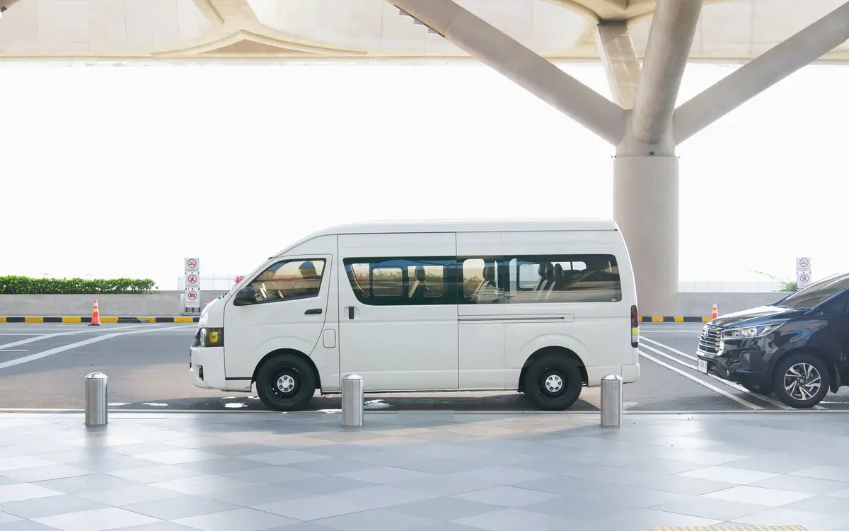 A white long wheelbase passenger van with tinted windows waiting in a kerbside drop off bay under an airport terminal canopy.