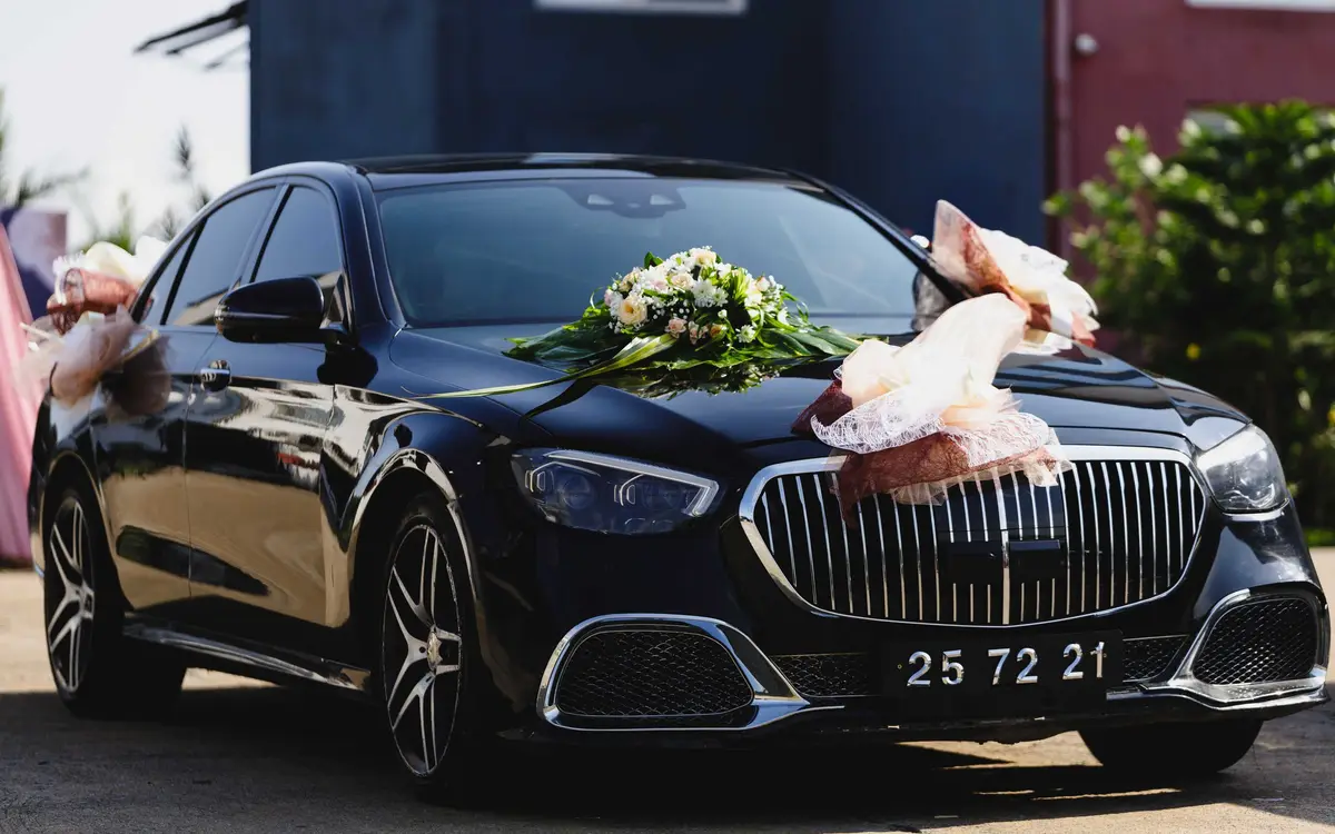 A polished black Mercedes Maybach saloon dressed for a wedding, with a cream and green floral arrangement across the bonnet and ribbon bows on the wing mirrors.